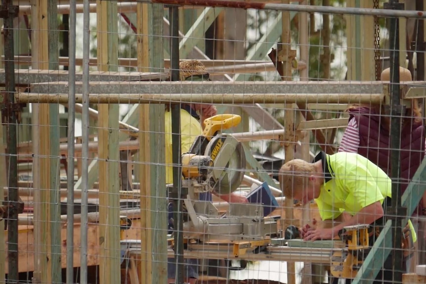 A workman wearing yellow works on construction site with wired fence and wooden frame of house.