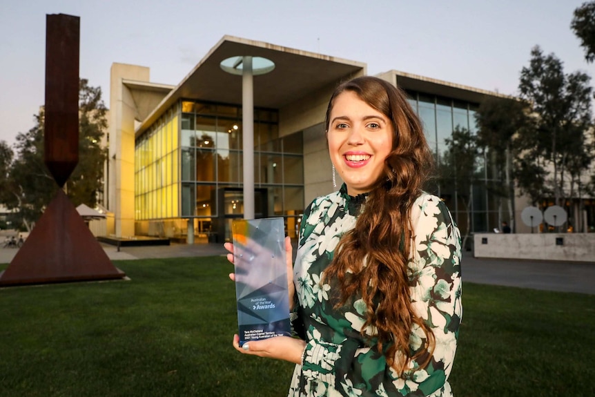 A young woman in a green and white dress stands before an architecturally designed building holding a glass trophy.