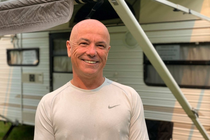 A bald man with a kind face smiling at the camera. He's standing in front of a caravan.