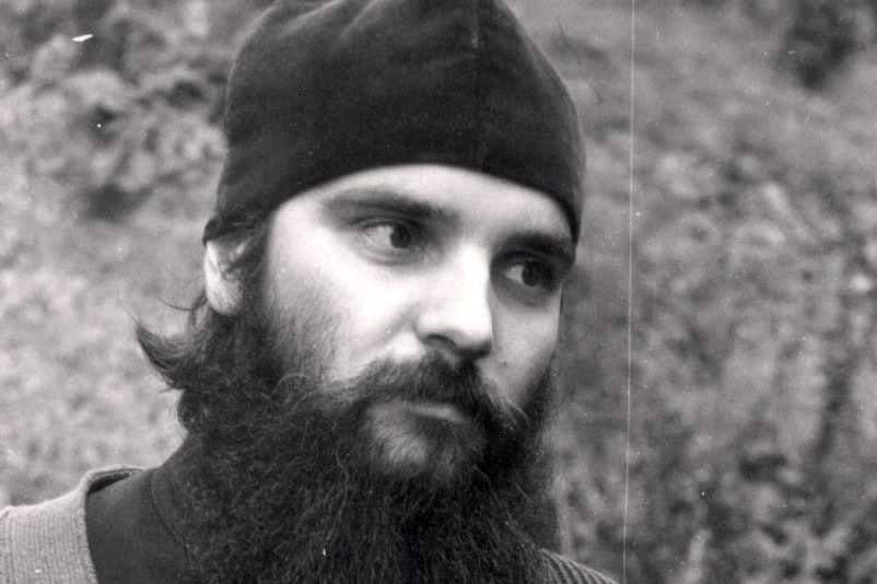 A black and white photo of a man outside with a long, black beard wearing a black beanie.