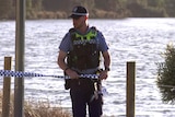 A police officer unspools crime scene tape with a rive behind him