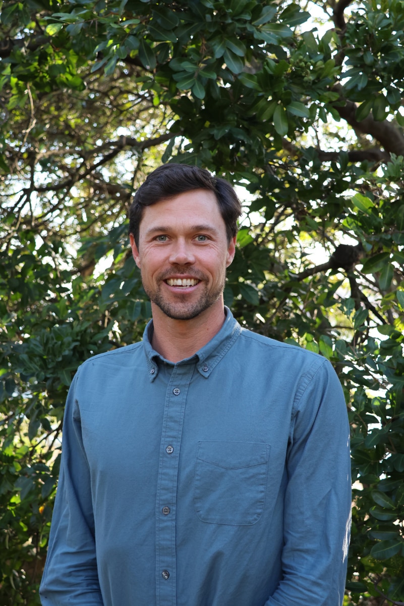 A man in a button-up shirt stands in front of trees and smiles at the camera