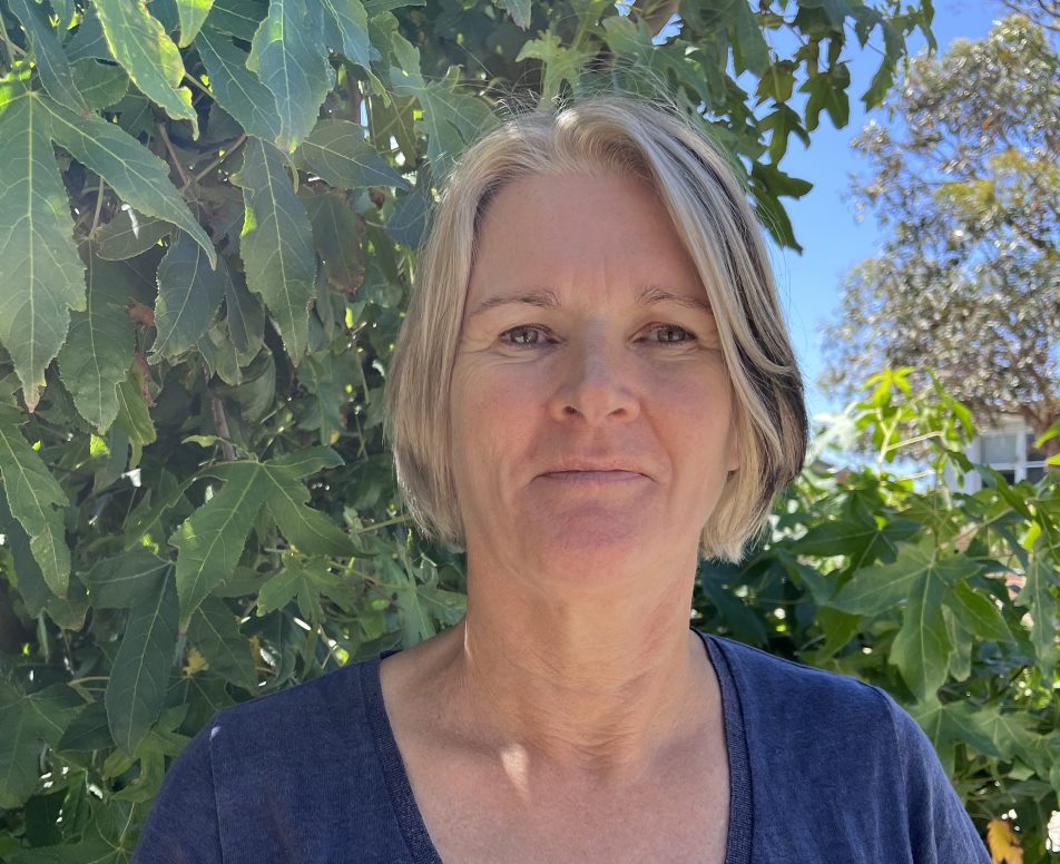 Woman with blonde hair poses in a headshot in front of green bushes