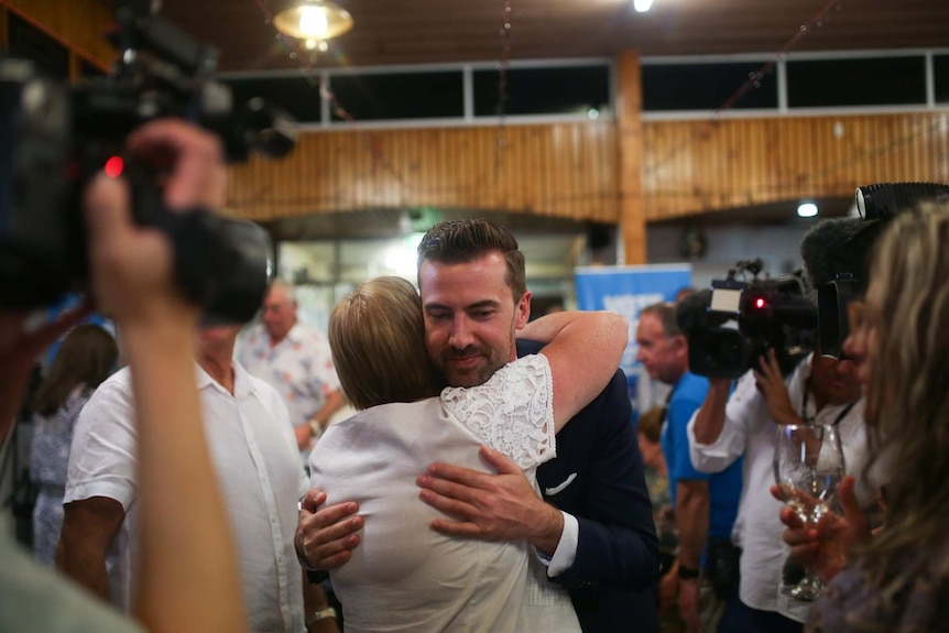 Zak Kirkup hugs a supporter with a half smile with multiple news cameras pointed at him