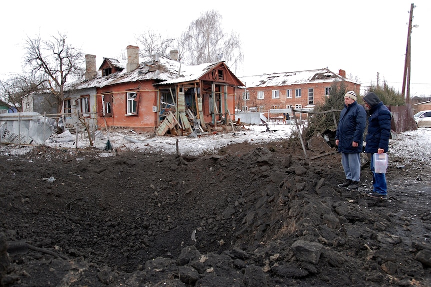 Two people stand next to a shell crater in front of a badly damaged house.