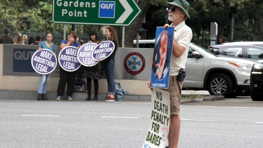 An anti-abortion protester holding placards stands across the road from pro-choice supporters, also holding placards.