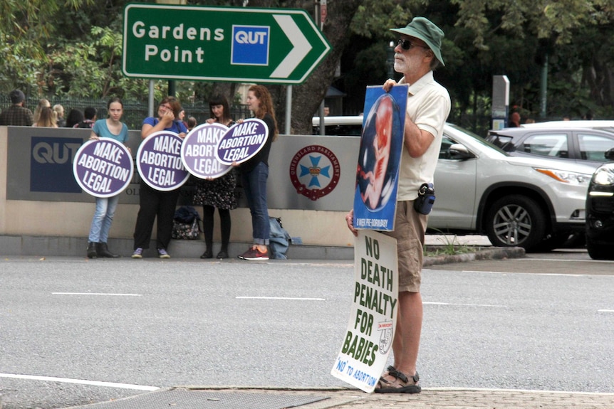 An anti-abortion protester holding placards stands across the road from pro-choice supporters, also holding placards.