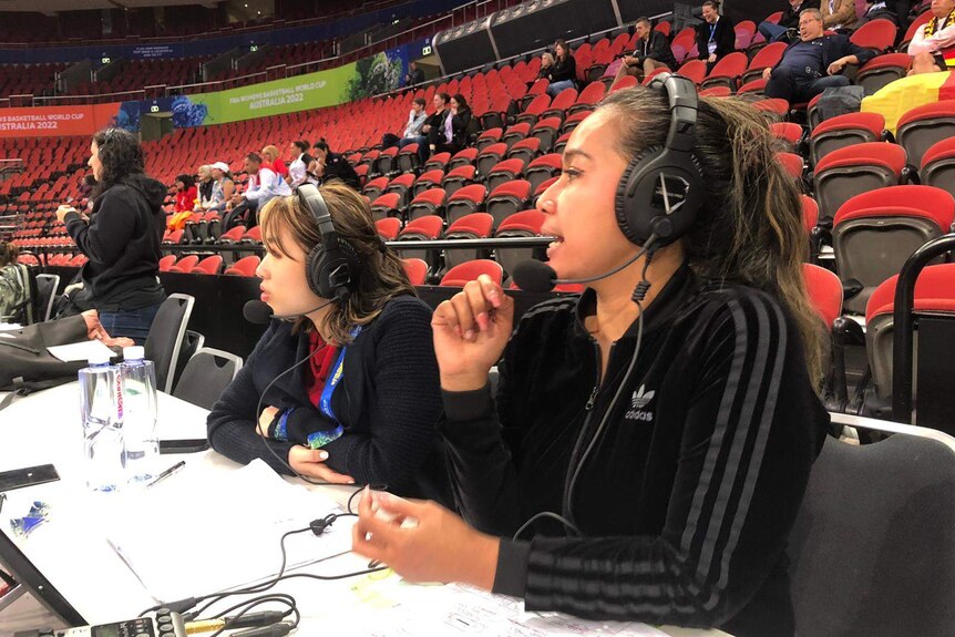 Two women sit at a commentators desk in a stadium wearing headphones and commentating on the game.