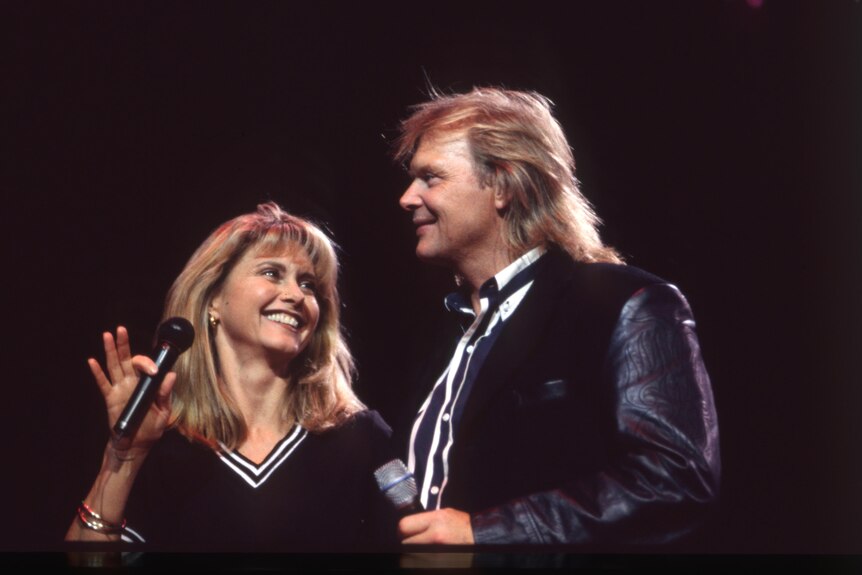 Olivia Newton-John holding a microphone on stage with John Farnham - both dressed in black with a touch of white stripes