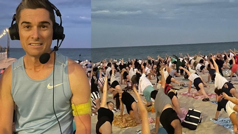 man in sleeveless blue shirt with headphones on sitting on wall with beach in background with hundreds doing yoga