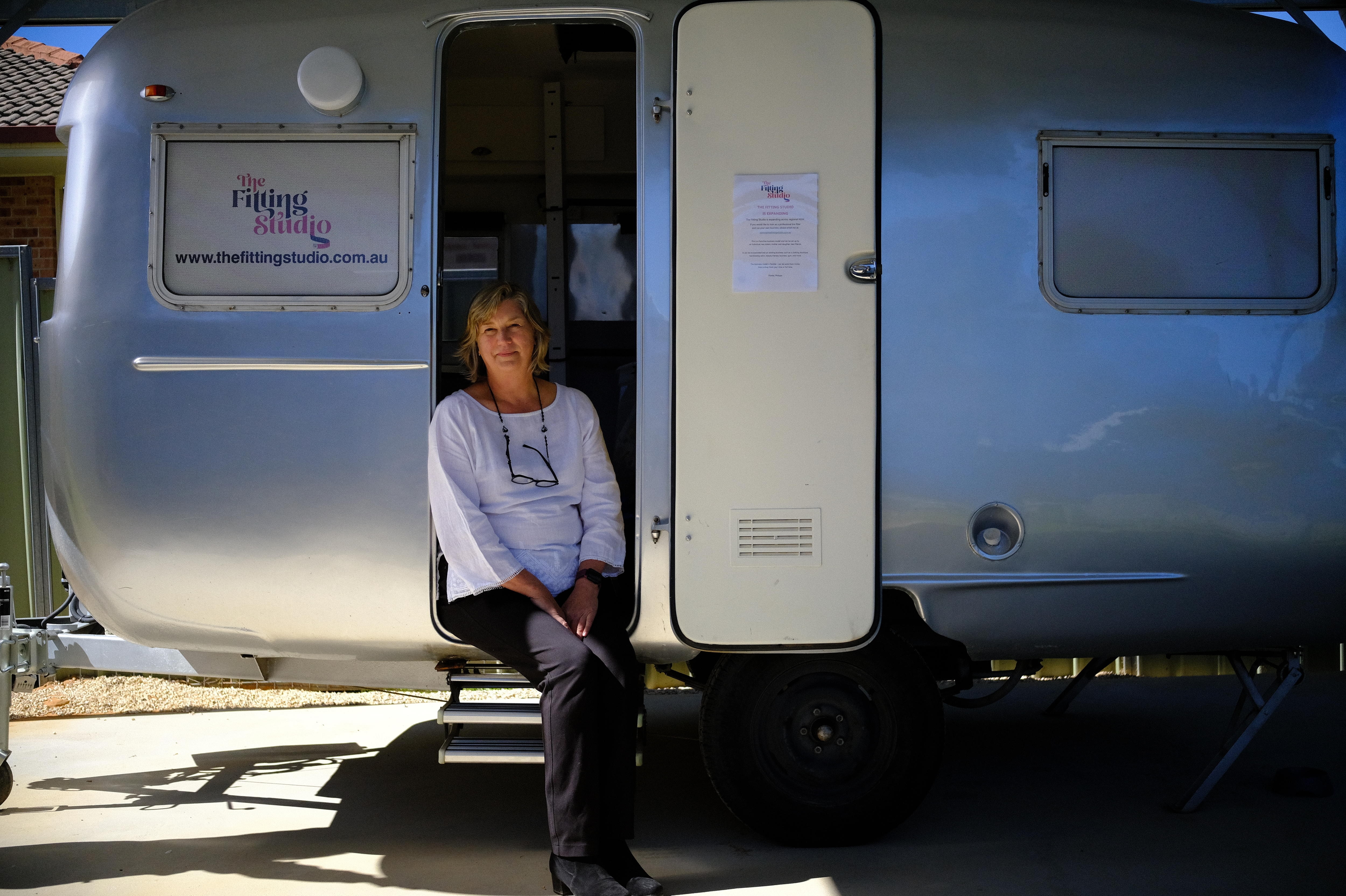 An older woman sits in the entry way of a caravan that has been fitted into a bra fitting studio.