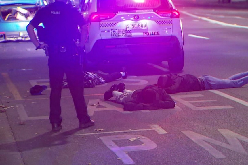 Three men lying on the ground face down in a bus lane with a police four-wheel drive and officer next to them