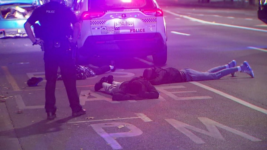 Three men lying on the ground face down in a bus lane with a police four-wheel drive and officer next to them