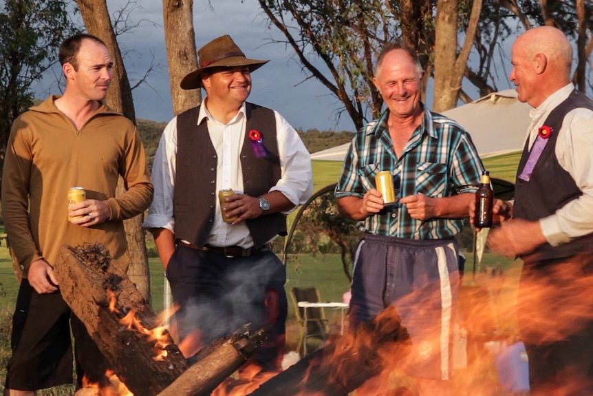 Four men drinking beer around a country campfire, wearing red remembrance poppies
