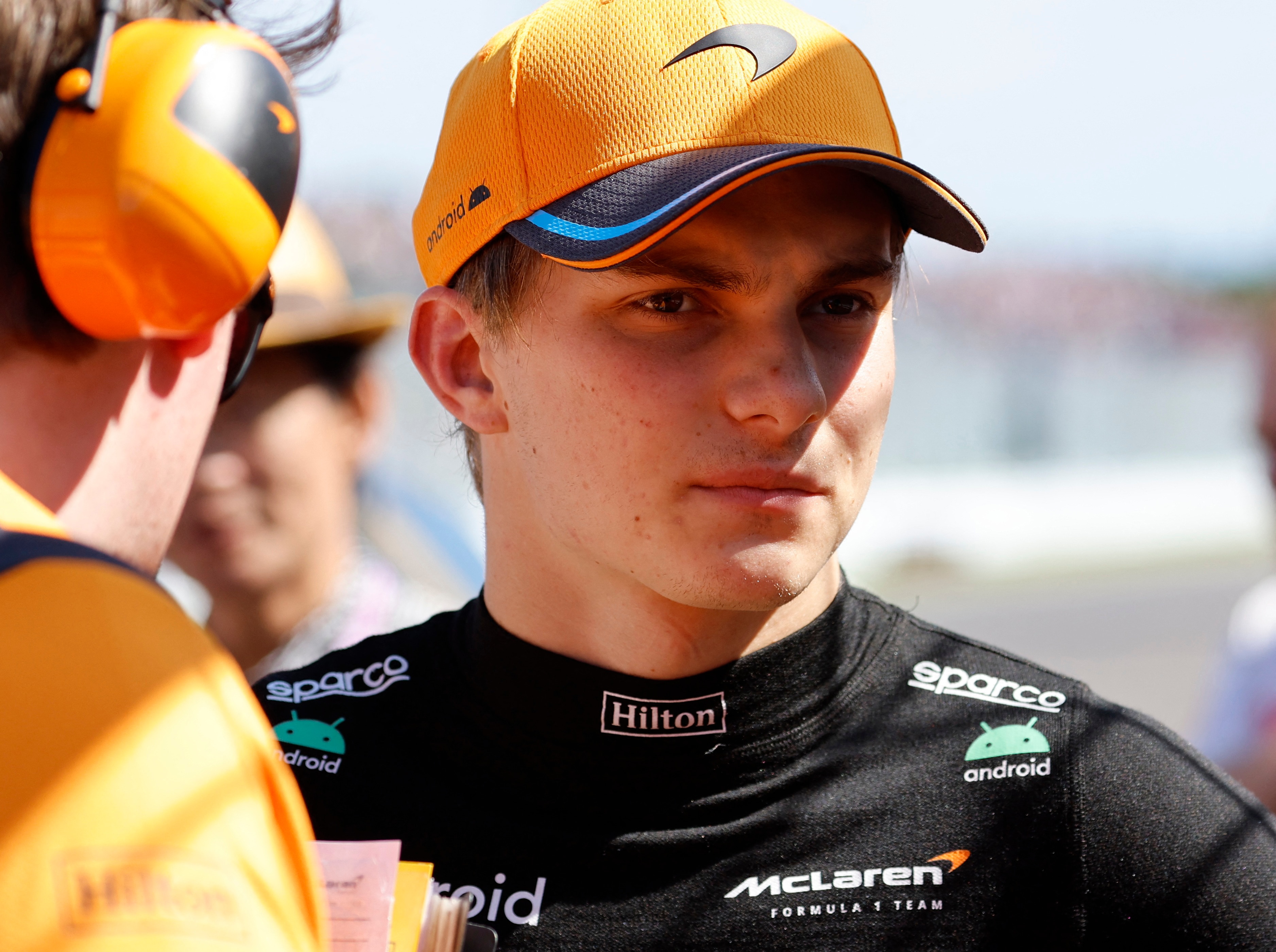 An F1 driver, out of the car, wearing a black shirt and orange cap, being talked to by a man with earmuffs