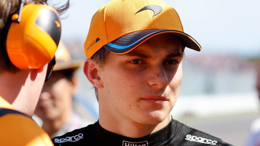 An F1 driver, out of the car, wearing a black shirt and orange cap, being talked to by a man with earmuffs