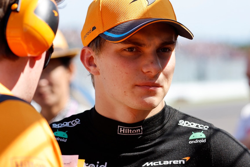 An F1 driver, out of the car, wearing a black shirt and orange cap, being talked to by a man with earmuffs