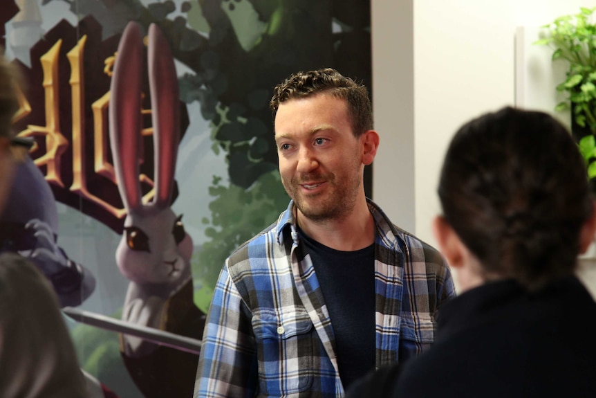 Blake Mizzi stands in front of a promotional poster for the video game Armello speaking to a group of people.