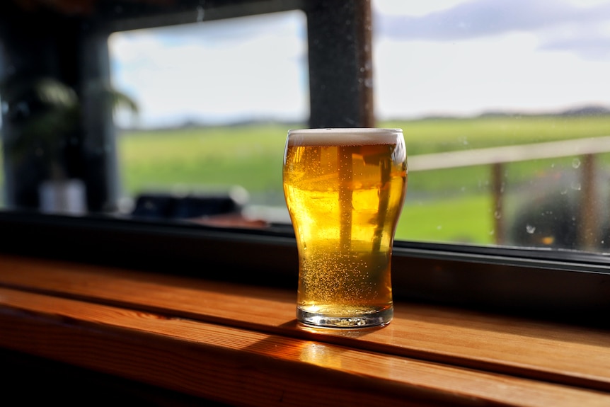 A glass of beer on a bench in front of a window with a pasture in the background