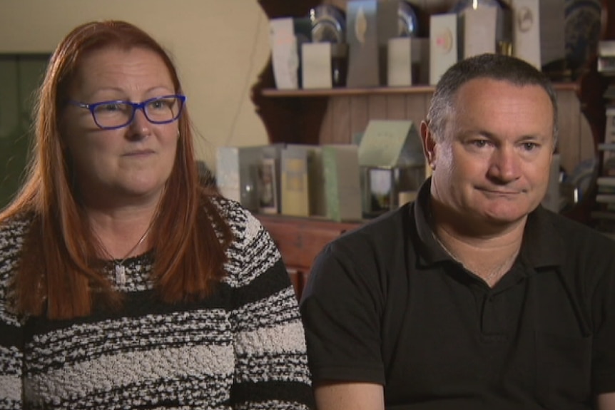 The mother and step father of Jason Challis sit in a lounge room looking at a camera with sad expressions.