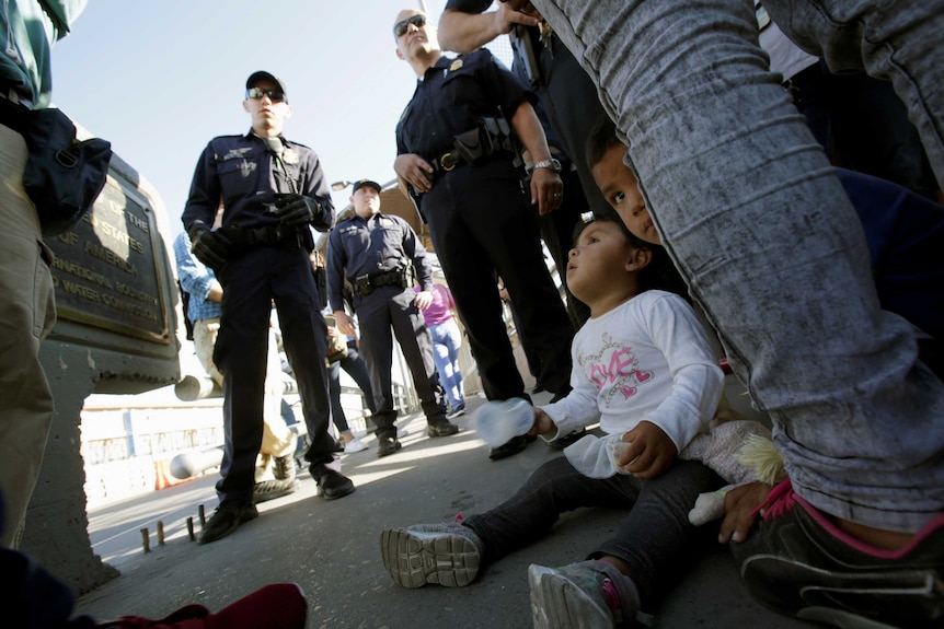 A photo from ground level shows children sitting on the ground surrounded by border protection authorities.