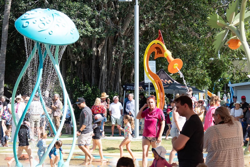 Dozens of adults and children playing at a waterpark, with a blue jellyfish structure spraying water onto a splash pad.