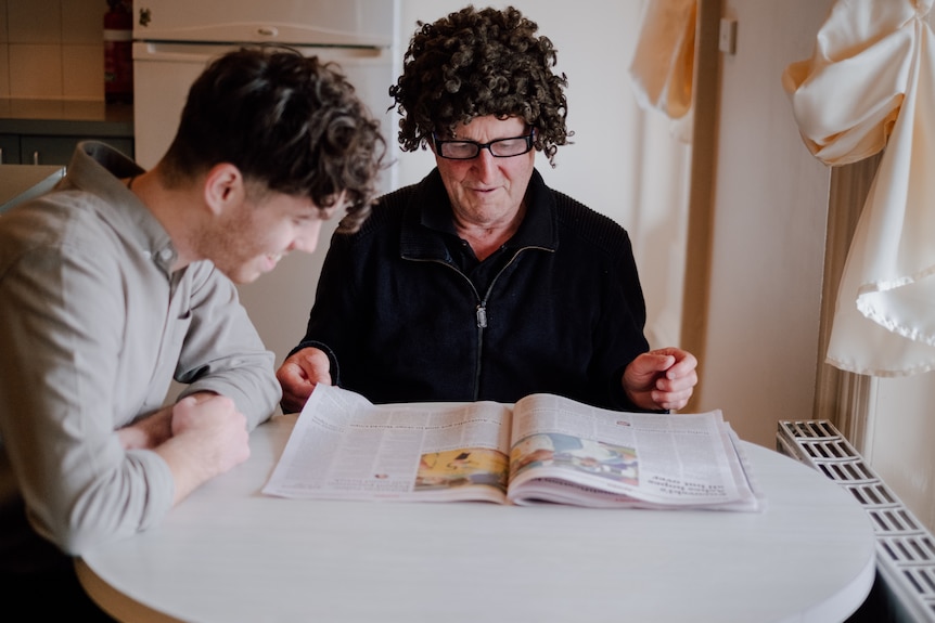 Two men sit at a table in a kitchen, smiling as they read a magazine together.