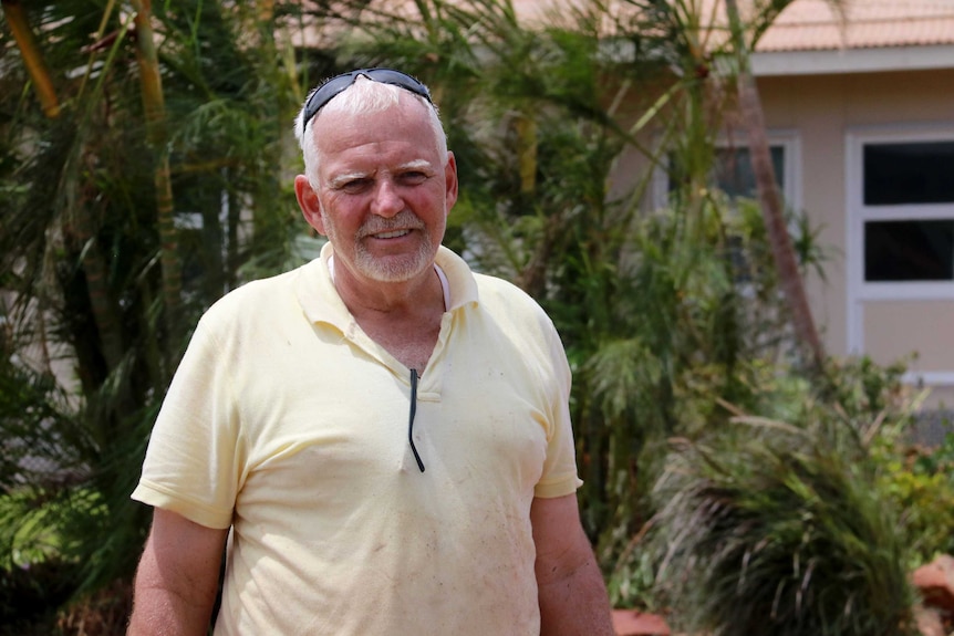 A man in a dirty shirt with a wind swept garden behind him.