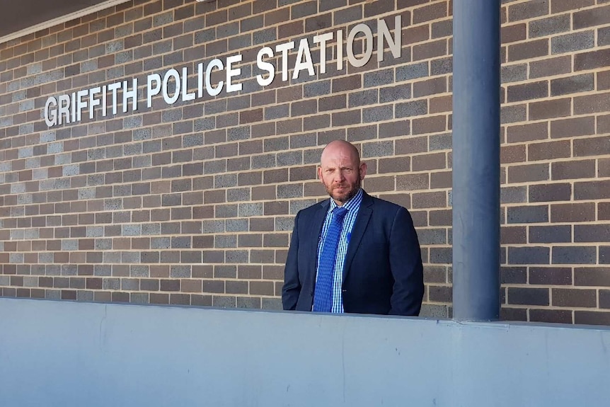 A bald, bearded, solidly-built police officer in a suit stands in front of Griffith Police Station.