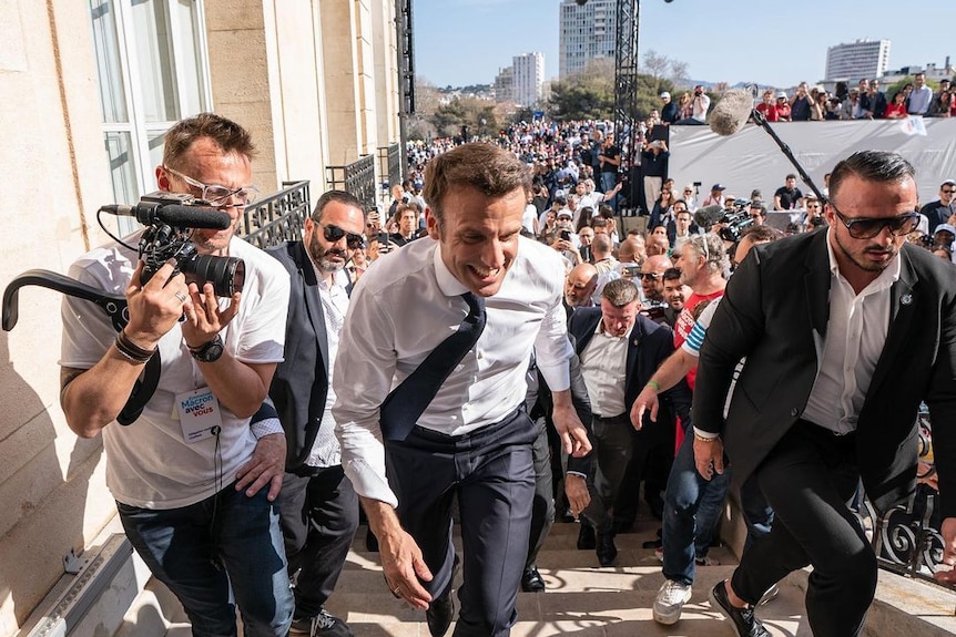 Emmanuel Macron smiles as he walks up the stairs closely followed by a cameraman.