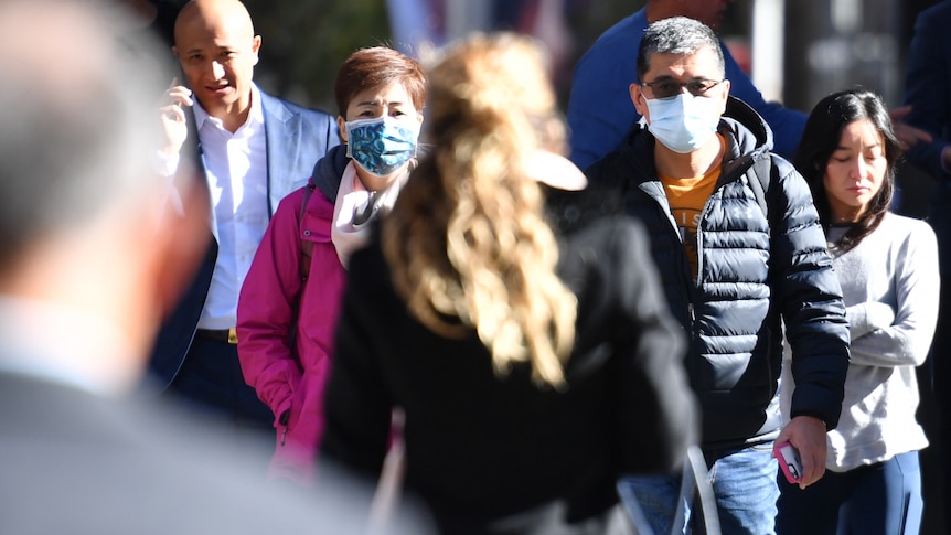 A group of people walk on a busy street, some wearing masks and some without.