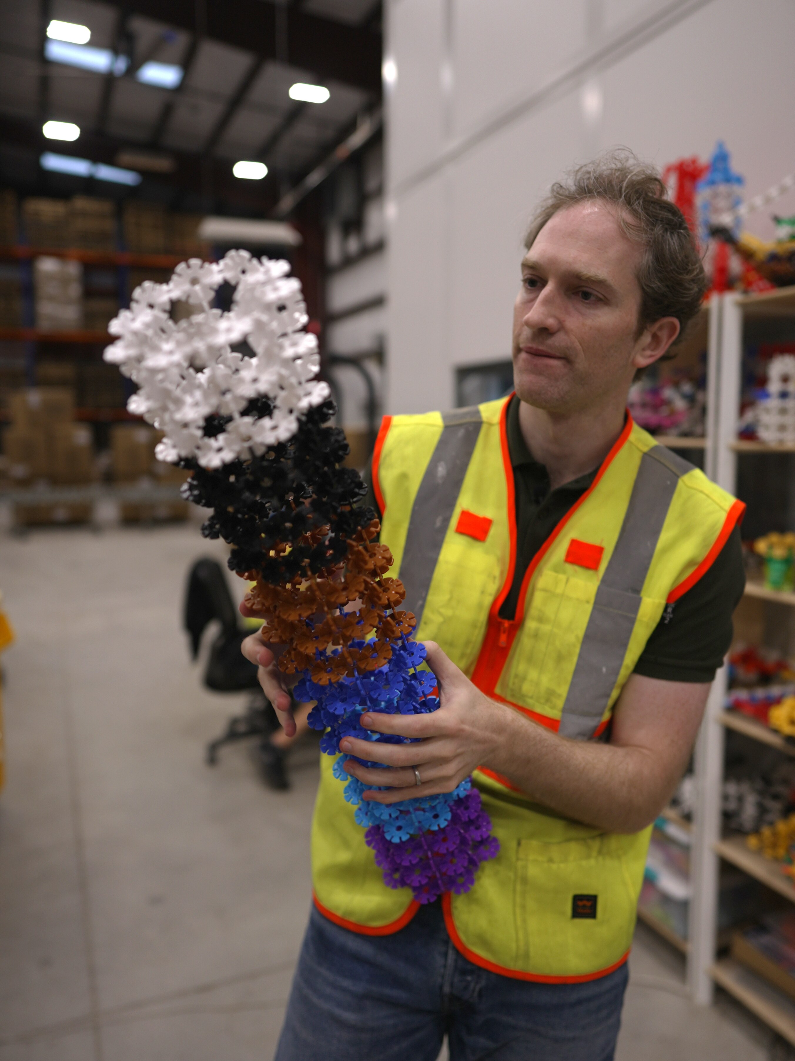 A man wearing a hi vis vest standing in a warehouse holds a building blocks toy. 