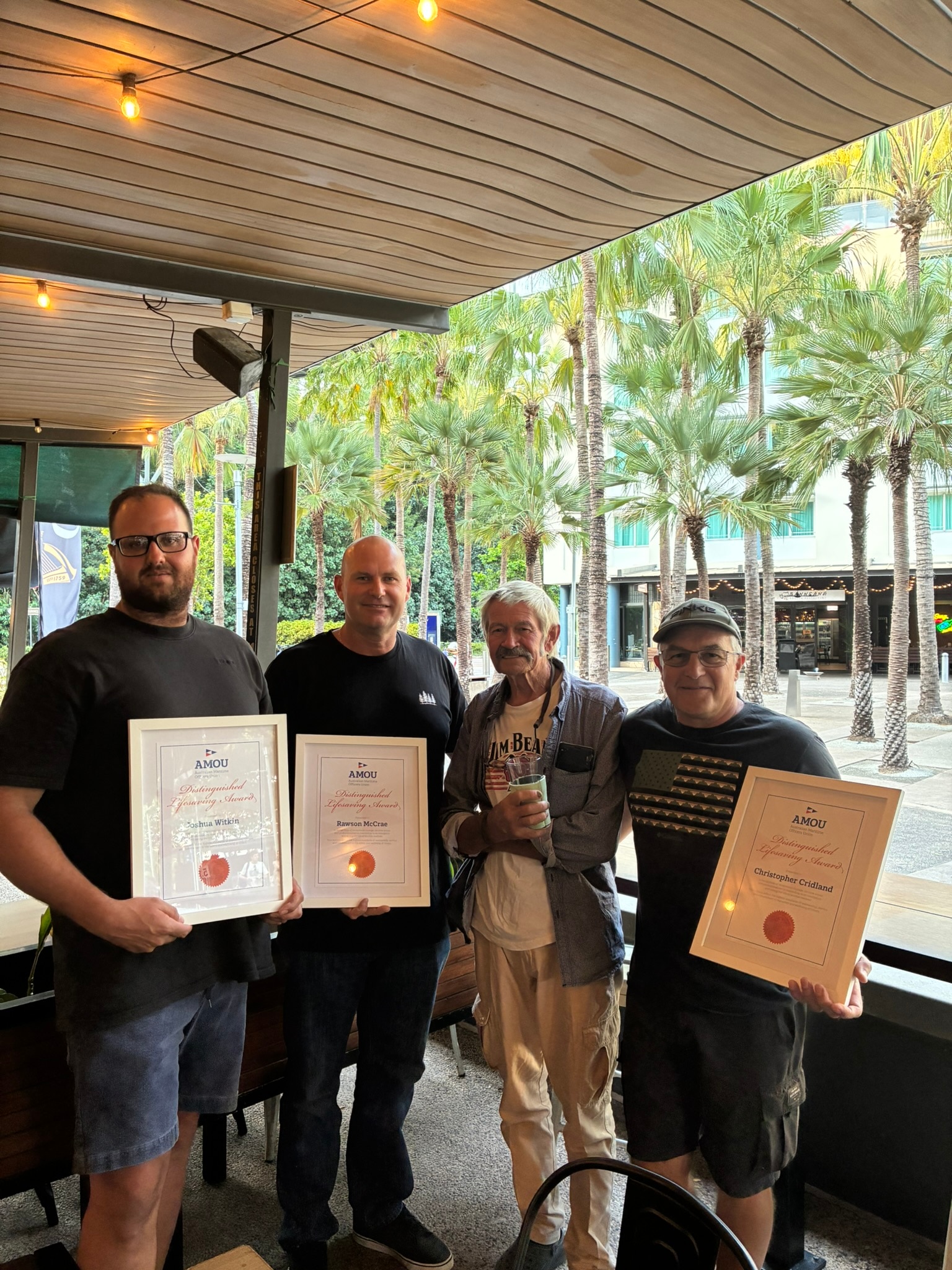 Four men stand in a line, three of them holding framed AMOU certificates