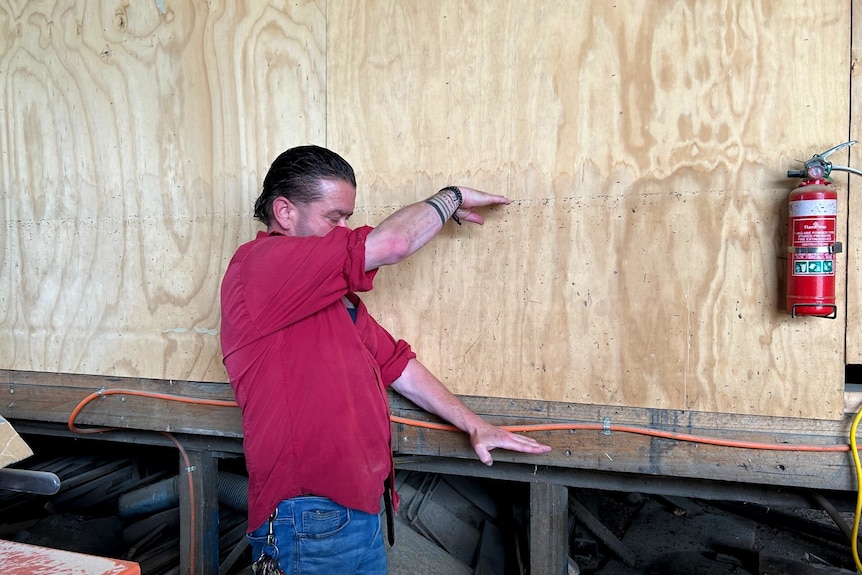 A man in a red shirt points to a timber wall