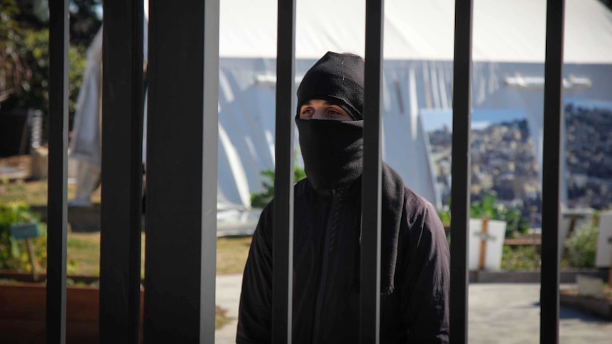 A masked man behind black bars.