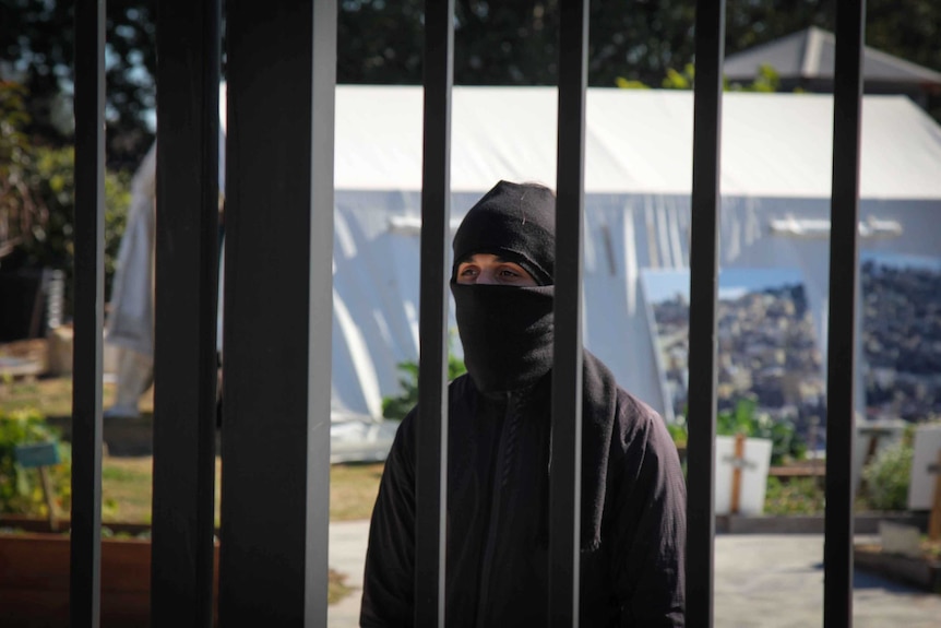A masked man behind black bars.