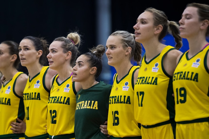 Seven women basketballers, wearing yellow Australia uniforms, have their arms around each other, signing the national anthem.