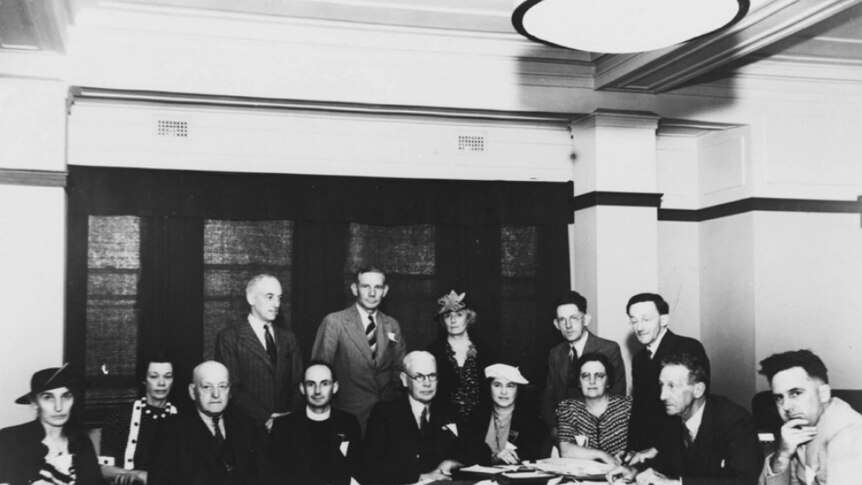 A group of men and women sit around a desk facing the Camera posing for a photo. Black and white.