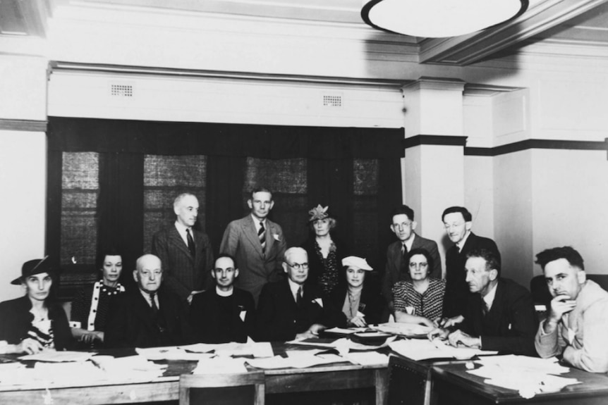 A group of men and women sit around a desk facing the Camera posing for a photo. Black and white.