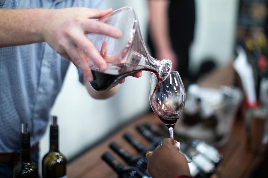 A hand is pouring red wine from a decanter into a wine glass being held by another person