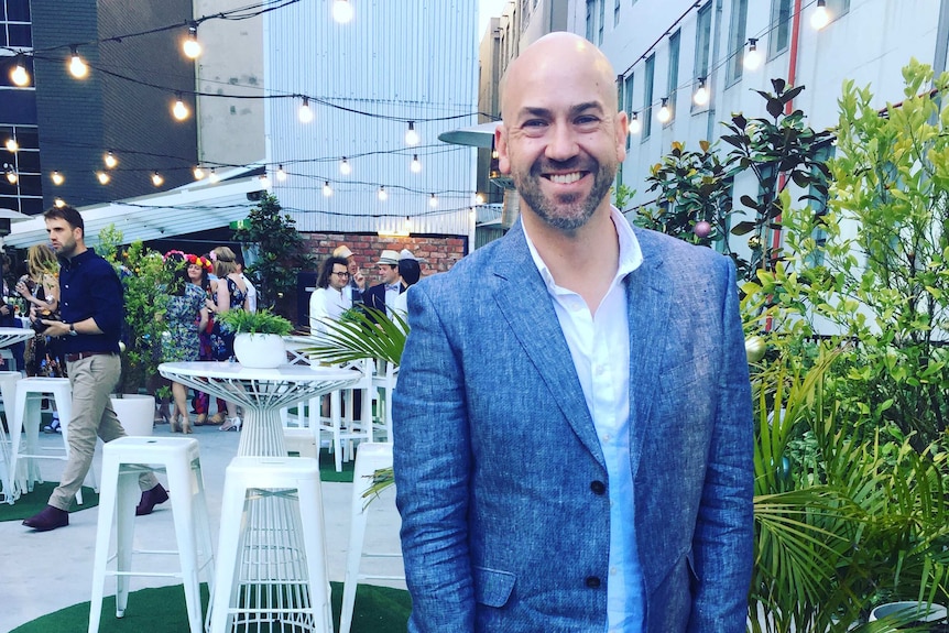 A man wearing a blazer stands smilingn in a busy courtyard or beer garden strung with fairy lights.