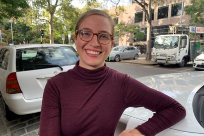 Anika Stobart leans up against a white car wearing a plum turtle neck top