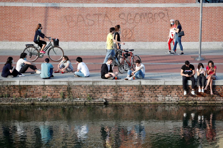 people with masks sit near, drink and ride past water and a brick wall in Milan