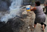 Woman throws water from white bucket onto smouldering ashes.