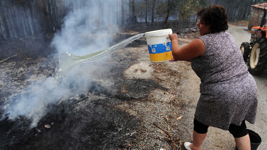 Woman throws water from white bucket onto smouldering ashes.