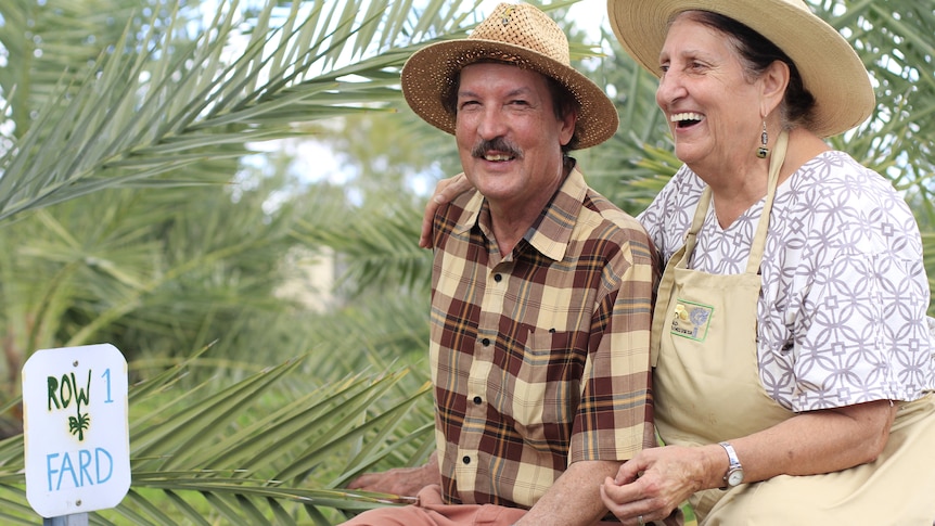 A smiling man and woman in wide brimmed hats smile beside a date palm.