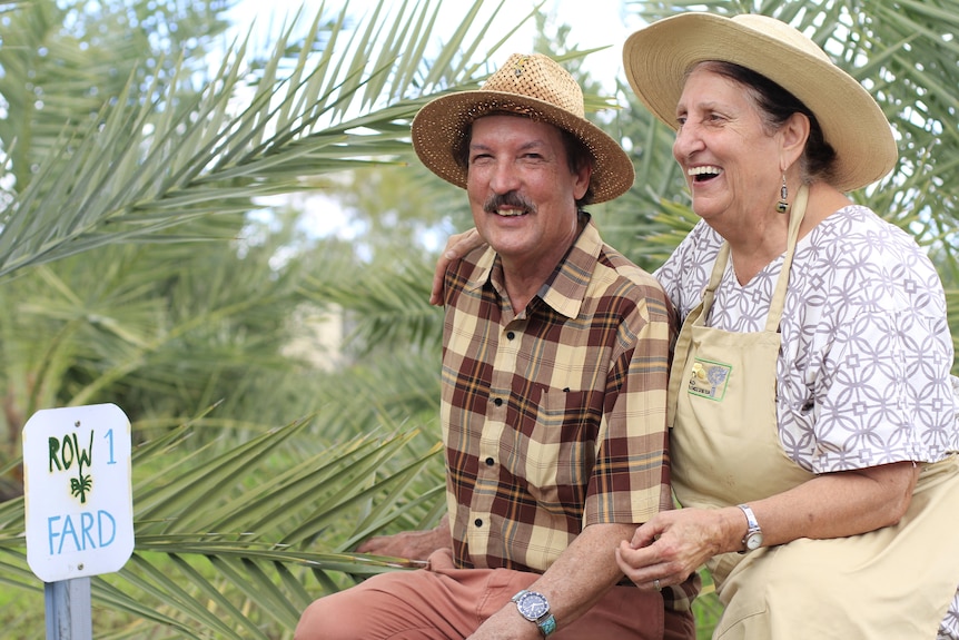 A smiling man and woman in wide brimmed hats smile beside a date palm.
