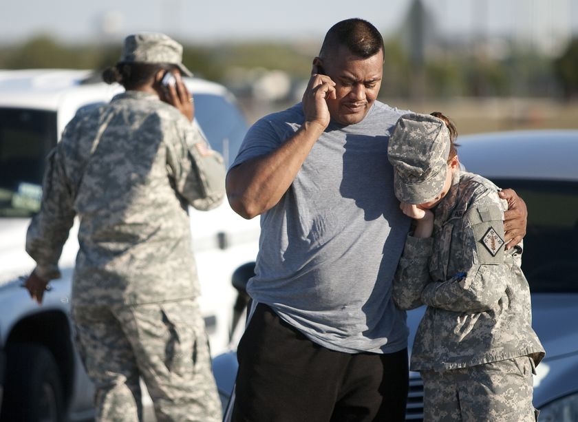 Man on a mobile phone with his arm around a young woman in uniform. Another female solider is on a mobile phone in background.