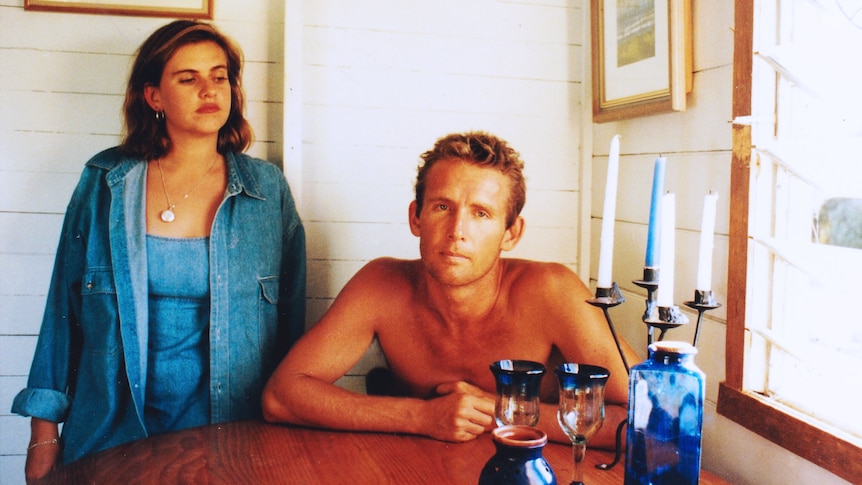 An old style photo of a shirtless blonde man and a woman wearing double denim. The man is sitting at the table