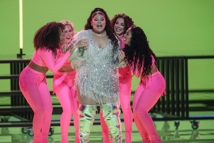 A woman stands in a sequin and tasselled dress surrounded by four other women in hot pink. The set behind is green.